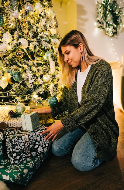 Happy caucasian woman puts present under Christmas tree while sitting on the floor at home.