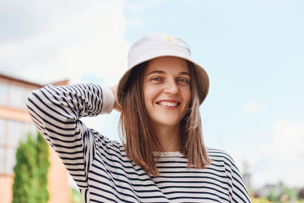 Happy Caucasian young woman wearing panama and striped shirt laughing looking at camera standing on street hipster girl posing outdoor summertime