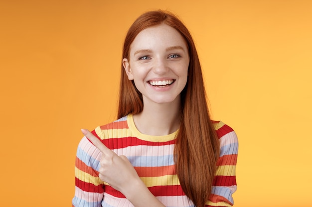 Happy charismatic smiling young ginger girl blue eyes sincere tender grin laughing broadly having fun discuss hilarious concert pointing upper left corner showing awesome product, orange background.