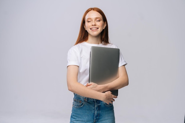 Happy charming young woman student holding laptop computer with closed eyes on isolated gray background. Pretty lady model with red hair emotionally showing facial expressions in studio, copy space.