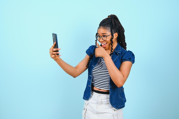 Happy cheerful Afro American woman looks at screen of smart phone enjoys online chatting types text message surfs social networks dressed casually poses against blue background Technology concept