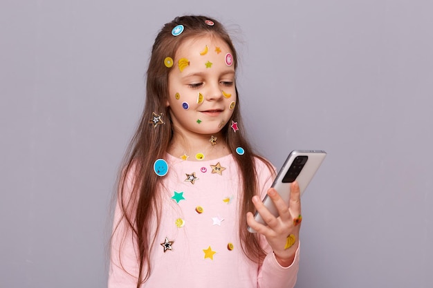 Happy cheerful little girl covered colorful kid's stickers standing isolated over gray background using smartphone for chatting with her friends in social networks