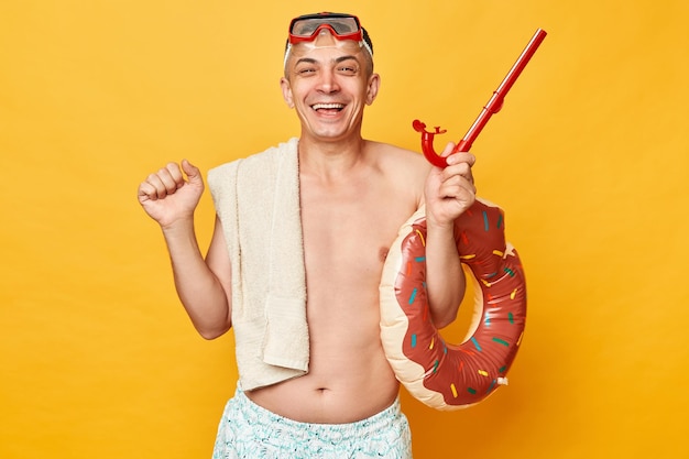 Happy cheerful man in diving goggles and snorkel holding donut rubber ring isolated on yellow background rejoicing his simmer activity clenched fists smiling