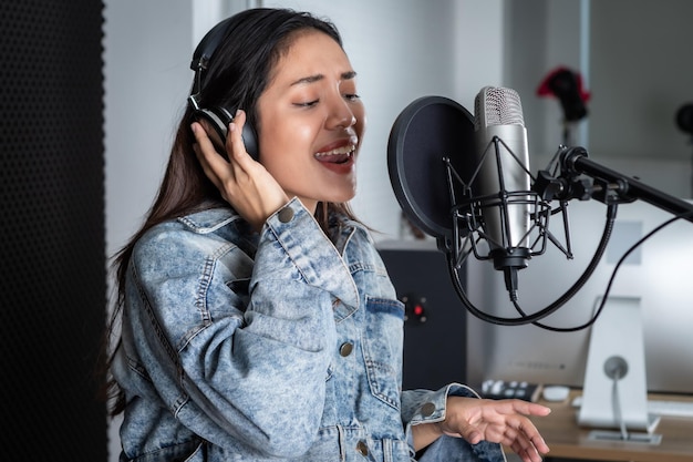 Happy cheerful pretty smiling of portrait of young Asian woman vocalist Wearing Headphones recording a song front of microphone in a professional studio