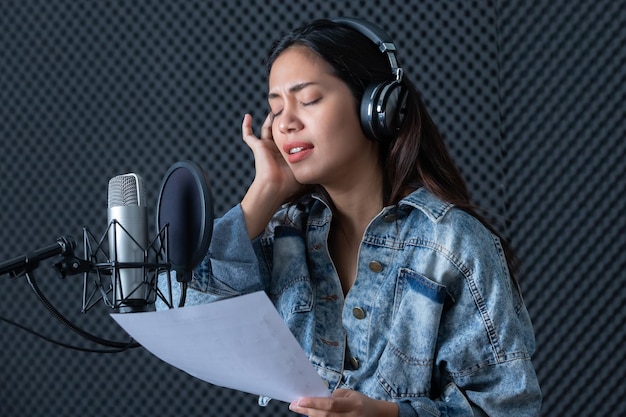 Happy cheerful pretty smiling of portrait of young Asian woman vocalist Wearing Headphones recording a song front of microphone in a professional studio