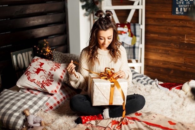 Happy cheerful young girl in a knitted vintage sweater opens a gift on a bed on Christmas Eve