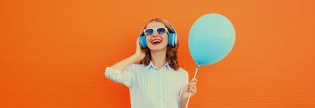 Photo happy cheerful young woman in headphones listening to music with blue balloon on orange background