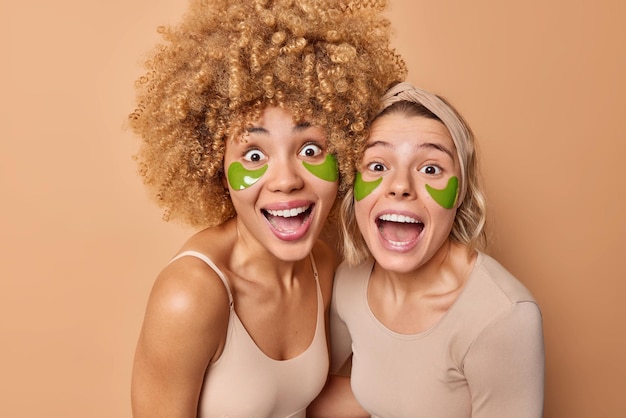 Happy cheerful young women look with amazed expressions keeps mouthes widely opened apply green hydrogel patches under eyes to remove wrinkles isolated over brown background Beauty concept