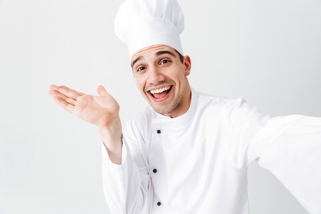 Happy chef cook wearing uniform standing isolated over white wall, taking a selfie