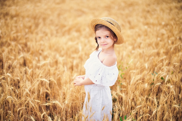 Happy child in autumn wheat field. Beautiful girl in white dress and straw hat have fun with playing, harvesting