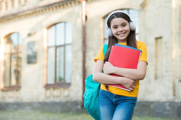 Happy child carry school bag and book wearing modern headphones for listening copy space