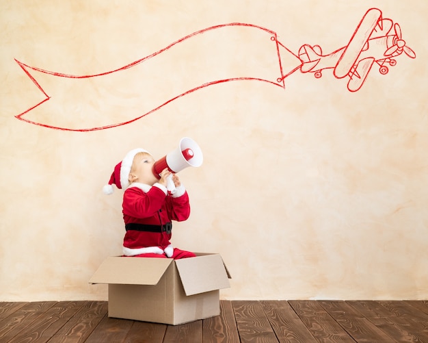 Photo happy child dressed santa claus costume playing at home. funny kid driving toy car and speaking by megaphone. christmas holiday concept