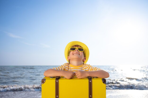 Happy child enjoying the sun against blue sky background Kid with suitcase having fun on the beach Summer vacation and holiday concept