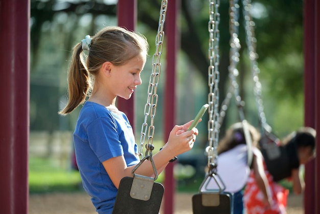 Happy child girl browsing her mobile phone sitting on swing in park during summer vacations