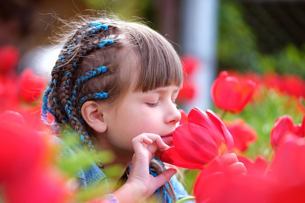 Happy child girl playing in summer garden enjoying sweet scent of red tulip flowers on sunny day