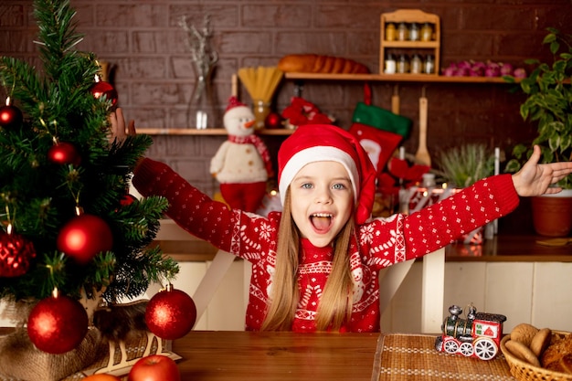 A happy child girl in a Santa hat in a dark kitchen at the Christmas tree with red balls rejoices and smiles the concept of new year and Christmas