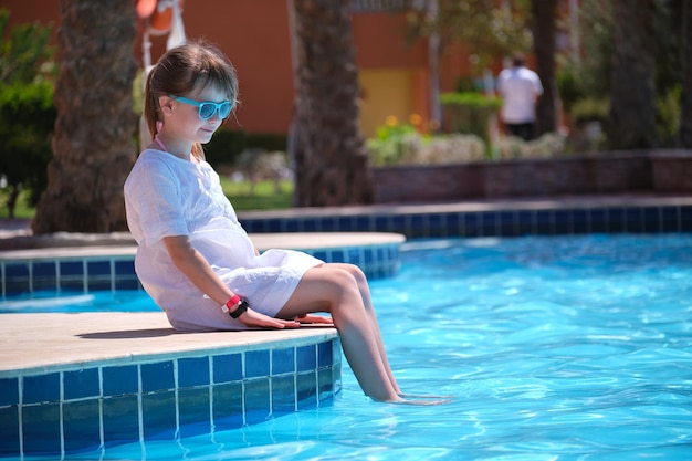 Happy child girl in white dress relaxing on swimming pool side on sunny summer day