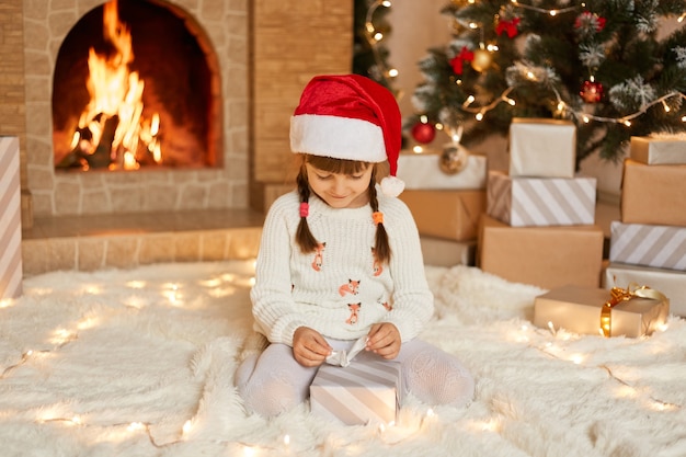 Happy child girl with Christmas present sitting on floor near xmas tree, looking at gift box with ribbon in her hands, wearing white jumper and red xmas hat, poses near fireplace.