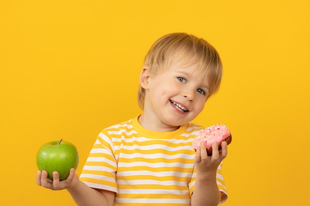 Happy child holding donut and apple against yellow wall.