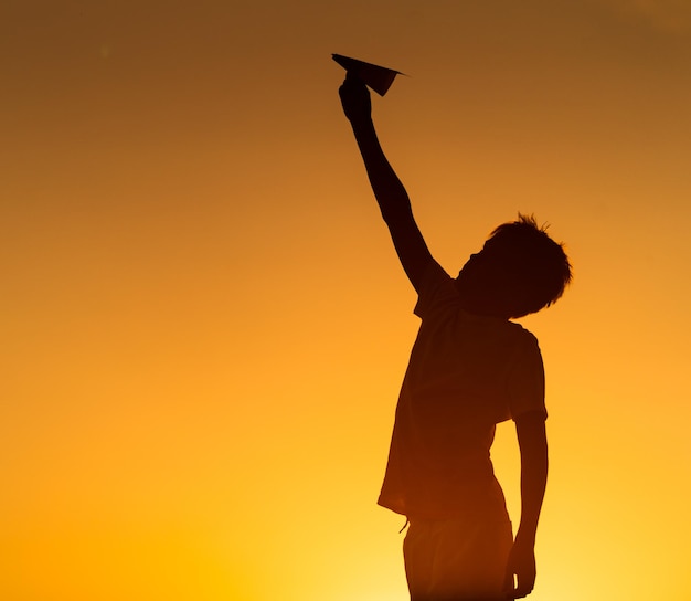 Happy child is playing with paper origami plane at orange sunset Vocation with children outdoors Silhuette against sunset