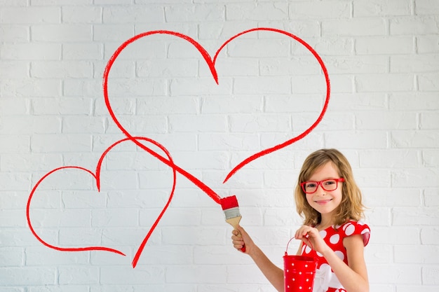 Happy child painting big red heart on the wall. Funny girl playing at home. Valentines day card. Renovation and design concept