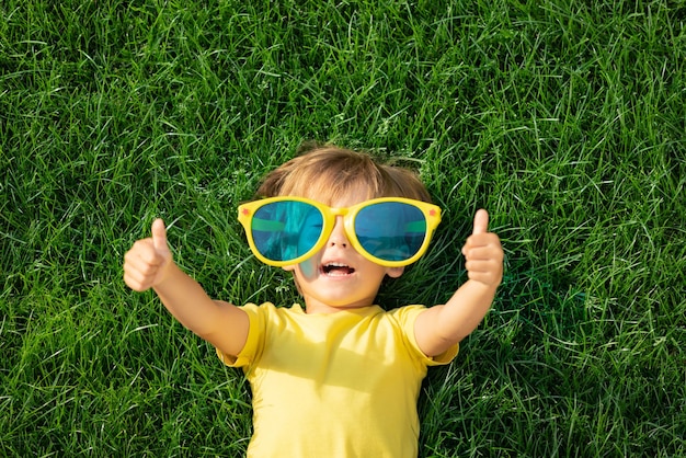 Happy child playing outdoor in spring park. Kid lying on green grass. Earth day and ecology concept. Boy showing thumbs up