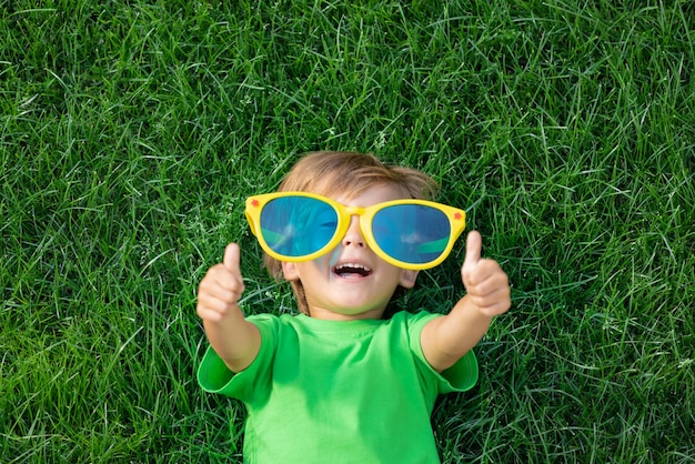 Happy child playing outdoor in spring park. Kid lying on green grass. Earth day and ecology concept. Boy showing thumbs up