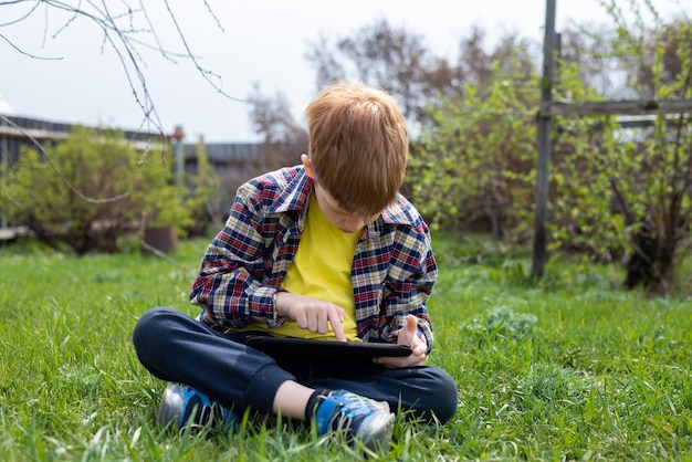 Happy child redhead boy playing on tablet sitting on green grass in the backyard in the village