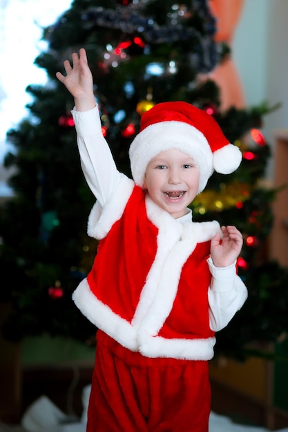 A happy child in a Santa Claus costume stands near the Christmas tree