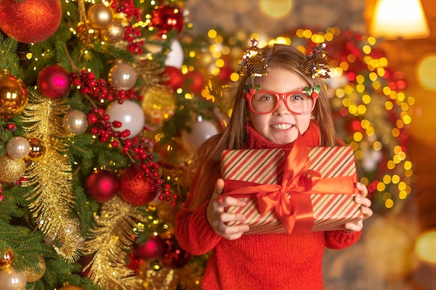 Happy child in a Santa red hat and in funny glasses is holding Christmas gift with smile. Christmas concept.