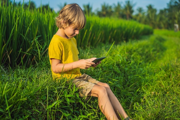 Happy child sitting on the field holding tablet Boy sitting on the grass on sunny day Home schooling or playing a tablet