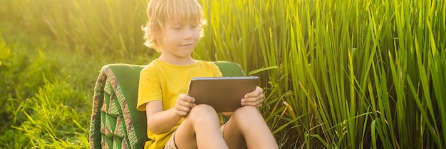 Happy child sitting on the field holding tablet boy sitting on the grass on sunny day home schooling