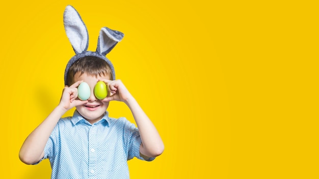 Happy child wearing bunny ears and holding colorful easter eggs in front of his eyes on a yellow background