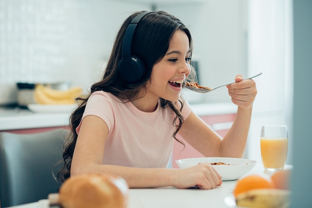 Happy child wearing headphones at home and smiling while eating a bowl of cornflakes with a glass of juice