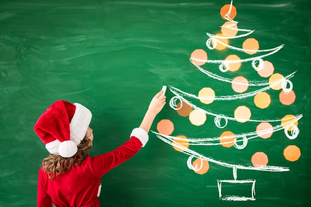 Photo happy child wearing santa claus draws on green blackboard