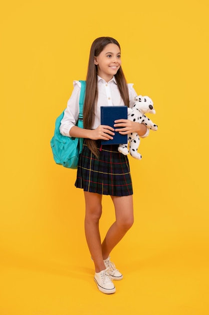 Happy child with backpack toy and book in school uniform full length on yellow background study