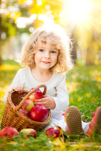 Happy child with basket of red apples in autumn park against golden sunny background