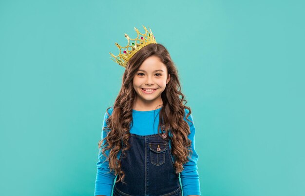 Happy child with curly hair in queen crown on blue background, selfishness.