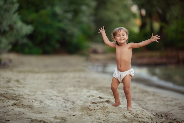 Happy childhood cheerful emotional child girl 1-2 years old plays on the sandy shore near the river. green trees grow behind her