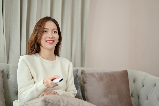 Happy and chill young Asian woman on the sofa holding a TV remote control and watching movie