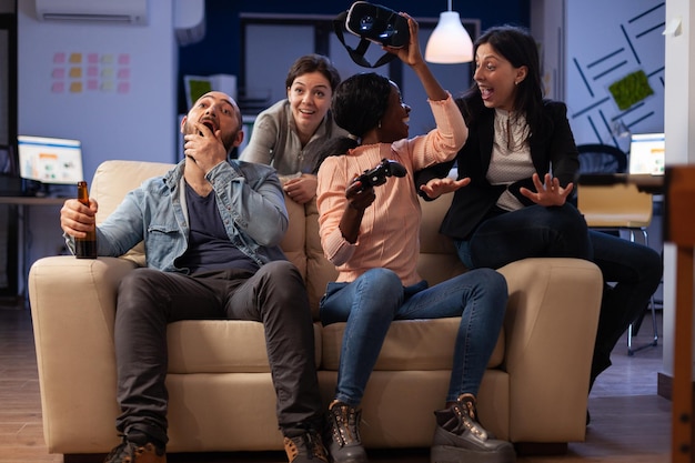 Happy colleagues using vr glasses on video games play with controller, having fun with drinks after work hours. Business people enjoying leisure activity with virtual reality goggles at office party.