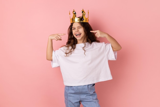 Happy confident smiling little girl standing in gold crown pointing at herself with pride