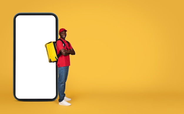 Happy confident young black delivery man in uniform with big backpack with crossed arms near phone