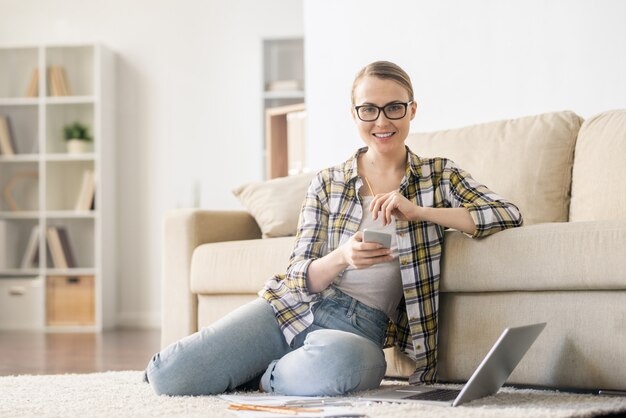 Happy confident young woman in glasses sitting on carpet in living room while using gadget and thinking about startup ideas