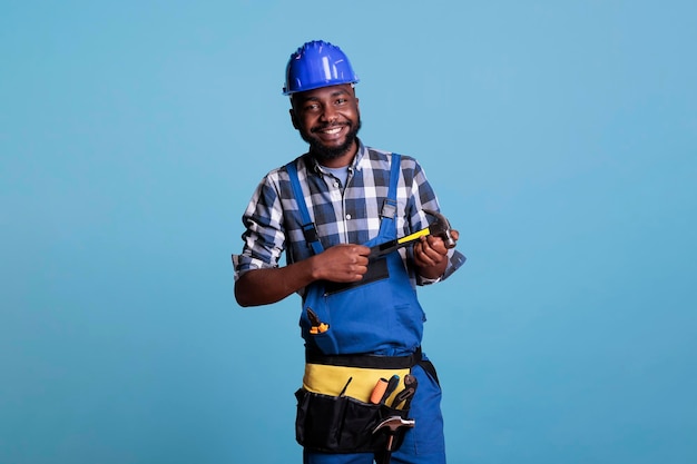 Happy construction worker smiling optimistic against blue isolated background. Studio photo of african american man wearing hard hat and tool belt holding hammer with both hands.