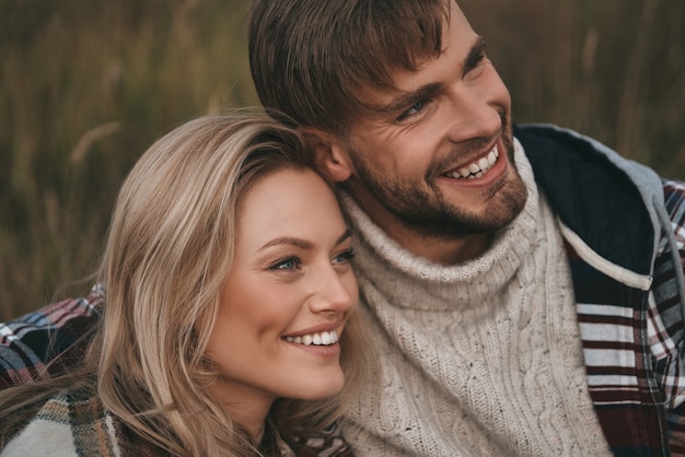 Happy couple.  Beautiful young couple embracing and looking away with smile while sitting on the field