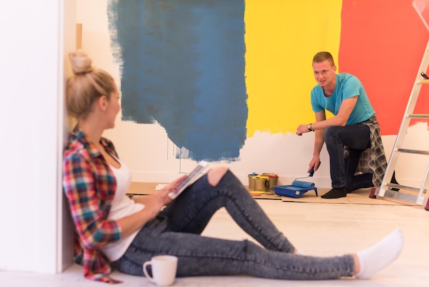 Photo happy couple doing home renovations, the man is painting the room and the woman is relaxing on the floor and connecting with a tablet