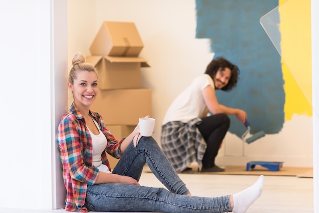 Photo happy couple doing home renovations, the man is painting the room and the woman is relaxing on the floor and drinking coffee