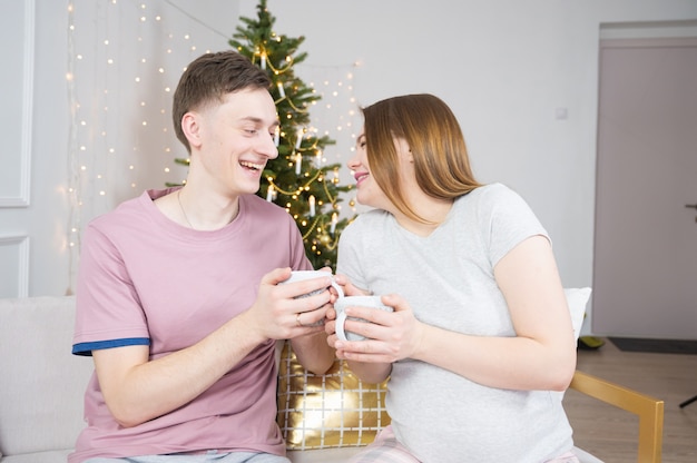 Happy couple drinking tea or coffee at home in evening over christmas tree lights on background. Holidays, leisure and people concept.