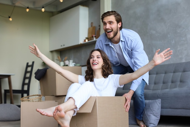 Happy couple is having fun with cardboard boxes in new house at moving day, woman riding sitting in cardboard box while man pushing it.